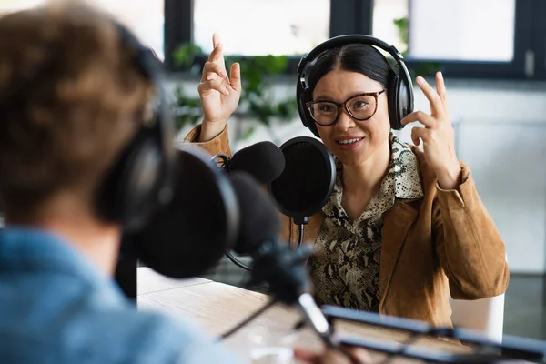 Asian announcer in glasses and headphones pointing up near blurred colleague — Stock Photo