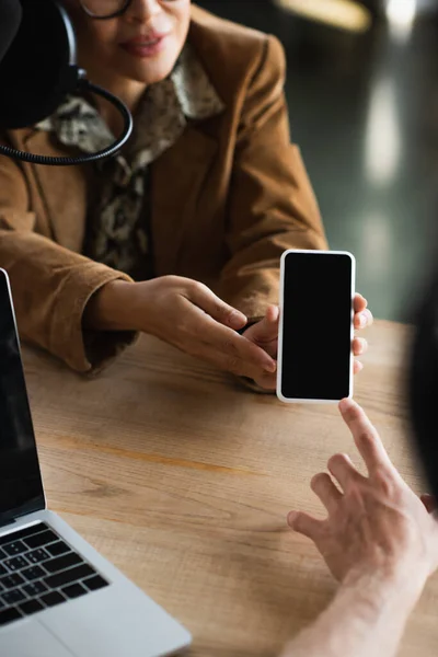 Cropped view of blurred announcer pointing at smartphone near colleague — Stock Photo