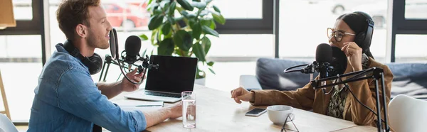 Interracial radio hosts recording podcast near gadgets and drinks in studio, banner — Stock Photo