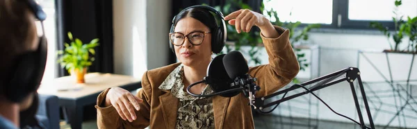 Asiatico radio host in occhiali e cuffie gesturing vicino sfocato collega in studio banner — Foto stock