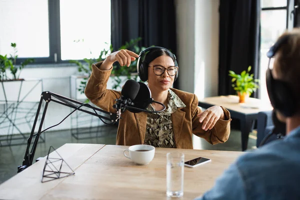 Asiático radio host en gafas y auriculares gesto cerca borrosa colega en estudio - foto de stock