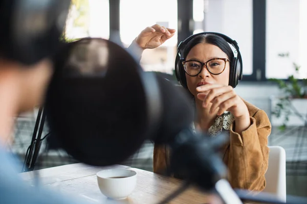 Asiático mujer en gafas y auriculares gesto cerca de taza de café y borrosa colega en difusión estudio - foto de stock