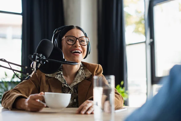 Felice asiatico radio host in occhiali e cuffie ridere mentre tiene la tazza in studio di trasmissione — Foto stock