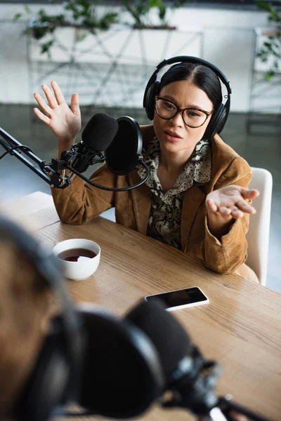 Der asiatische Radiomoderator mit Brille und Kopfhörer gestikuliert beim Podcast in der Nähe einer Tasse Kaffee und verschwommener Kollege — Stockfoto