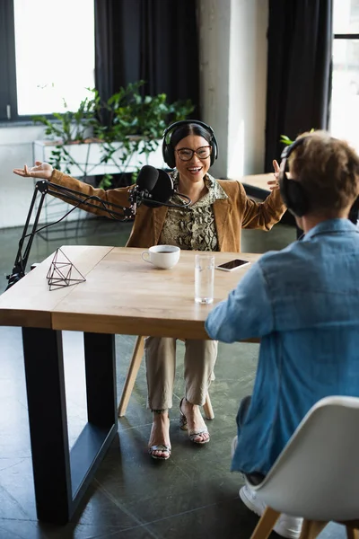 Feliz asiático radio host en gafas y auriculares gesto cerca de bebidas y borrosa colega durante podcast - foto de stock