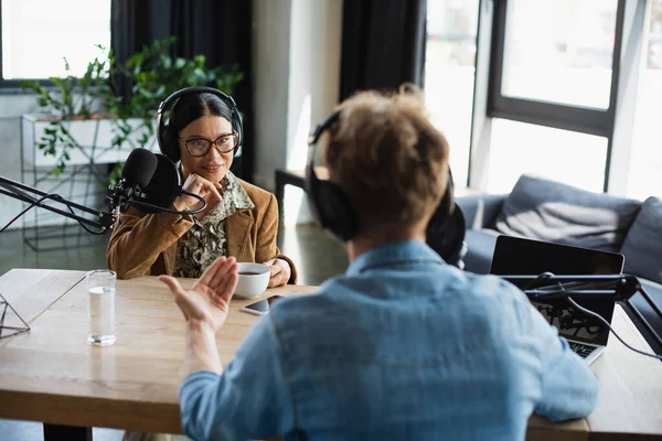 Happy asian radio host in eyeglasses and headphones looking at blurred colleague during podcast — Stock Photo