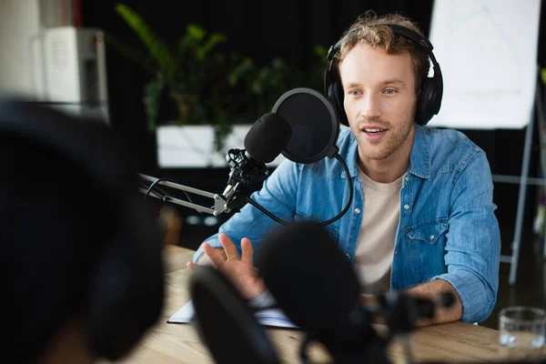 Anfitrión de radio en los auriculares mirando colega borrosa y gesto durante el podcast - foto de stock