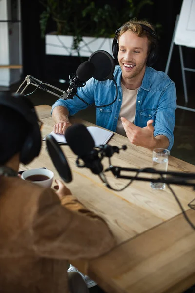 Anfitrión de radio alegre en los auriculares mirando colega borrosa y gesto durante el podcast - foto de stock