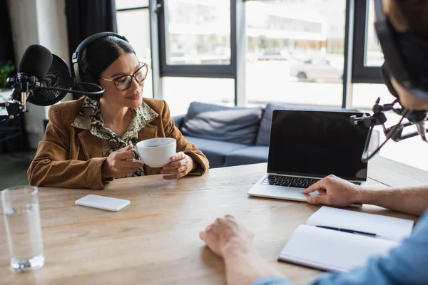 Asian radio host in eyeglasses and headphones looking at laptop during podcast — Stock Photo