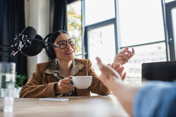Alegre asiático radio host en gafas y auriculares hablando con colega y celebración taza durante podcast - foto de stock