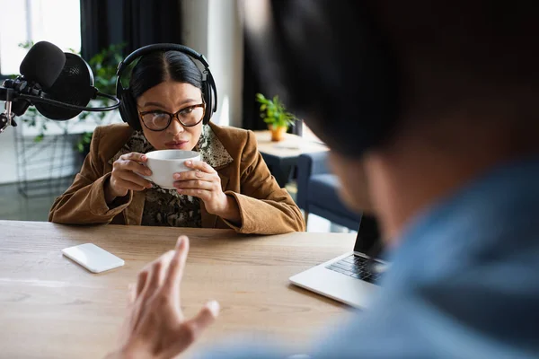 Asiático radio host en gafas y auriculares beber café cerca borrosa colega - foto de stock