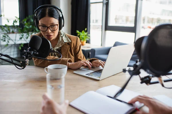 Asiático radio host en gafas y auriculares usando portátil y hablando en micrófono durante podcast - foto de stock