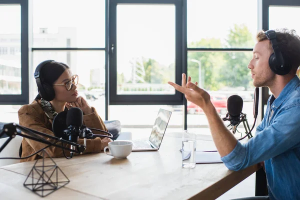 Interracial radio hosts recording podcast near laptop and drinks in studio — Stock Photo