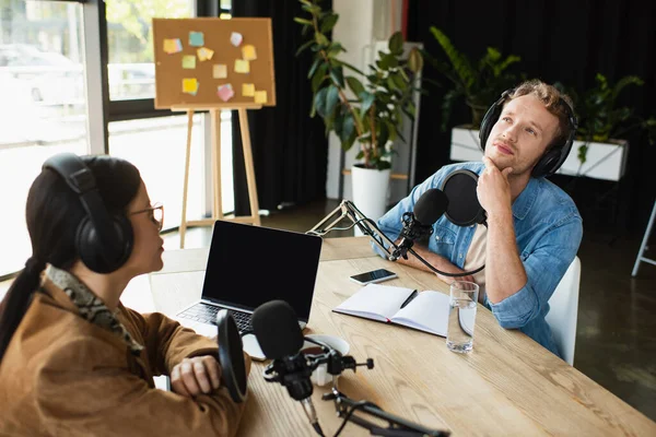 Pensive interracial radio hosts recording podcast near gadgets and beverages in studio — Stock Photo