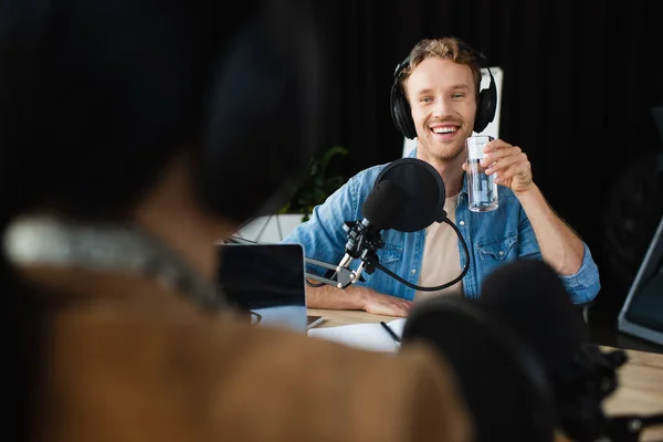 Anfitrión de radio feliz en los auriculares mirando colega borrosa y sosteniendo un vaso de agua durante el podcast - foto de stock