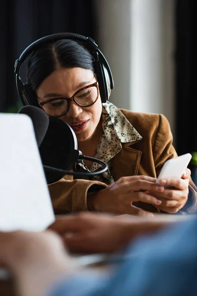 Asian radio host in headphones using smartphone near blurred colleague typing on laptop — Stock Photo