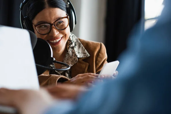 Щасливий азіатський радіоведучий в навушниках, використовуючи смартфон біля розмитого колеги, що друкує на ноутбуці — стокове фото