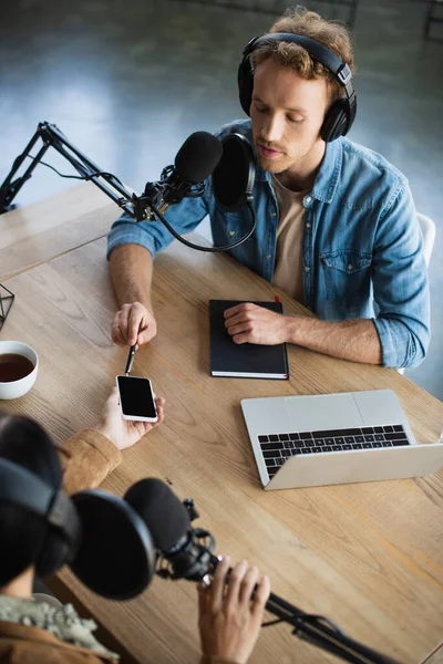 High angle view of radio host talking in microphone and pointing with pen at smartphone during podcast — Stock Photo