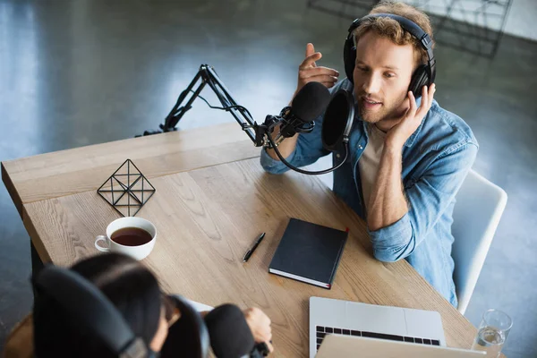 High angle view of radio host talking in microphone and gesturing near laptop and colleague during podcast — Stock Photo