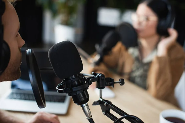 Radio host in wireless headphones talking in microphone near blurred asian colleague — Stock Photo