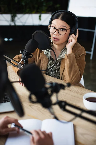 Sorpreso asiatico radio host in occhiali e cuffie parlando e guardando offuscata collega — Foto stock