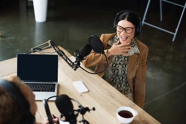 High angle view of joyful asian radio host in glasses and headphones laughing while looking at blurred colleague — Stock Photo