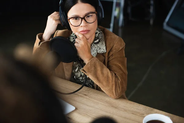 Thoughtful asian radio host in glasses and headphones looking at blurred colleague — Stock Photo