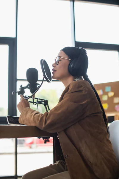 Вид збоку азіатського радіо хоста в окулярах і навушниках, що розмовляють, роблячи подкаст в студії — стокове фото