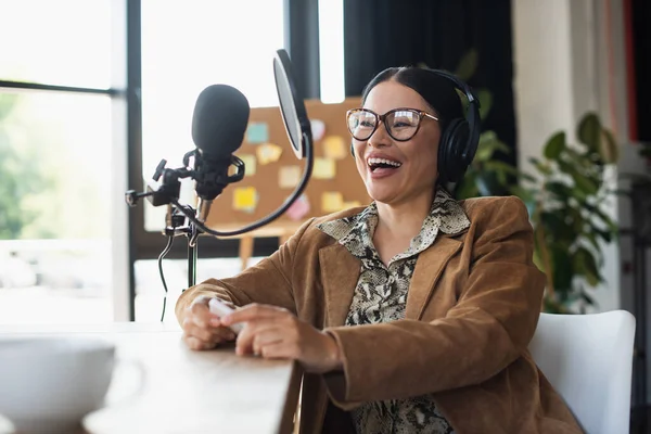 Позитивний азіатський радіоведучий в окулярах і навушниках тримає смартфон під час сміху під час подкасту — стокове фото