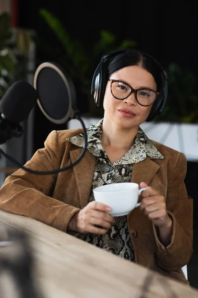 Asiático radio host en gafas y auriculares celebración taza de café y mirando a cámara - foto de stock