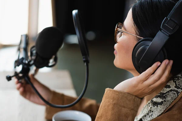 Hôte radio asiatique dans des lunettes et écouteurs toucher bras ciseaux avec micro pendant podcast — Photo de stock