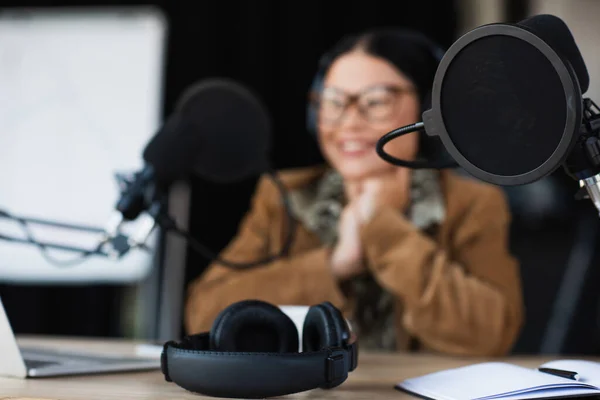 Modern microphone with pop filter near wireless headphones and blurred asian radio host — Stock Photo