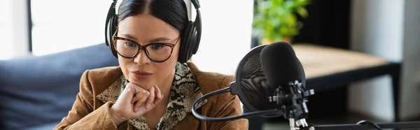 Pensativo asiático radio host en gafas y auriculares cerca de micrófono con filtro pop durante podcast, bandera - foto de stock