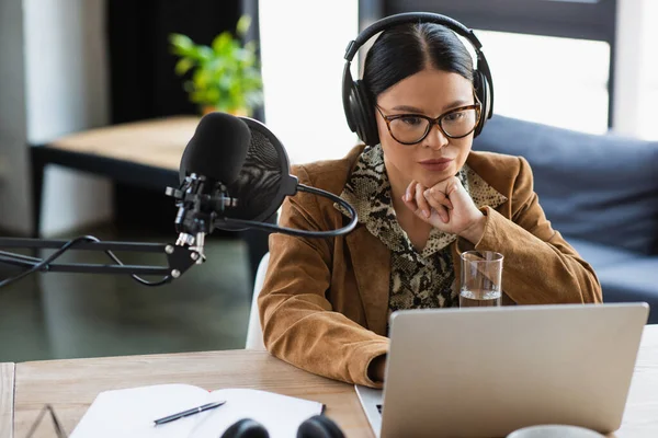 Pensive asian radio host in eyeglasses and headphones using laptop near near microphone with pop filter — Stock Photo