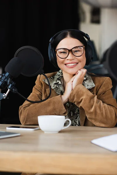 Feliz asiático radio host en gafas y auriculares sonriendo cerca de taza y smartphone en escritorio - foto de stock