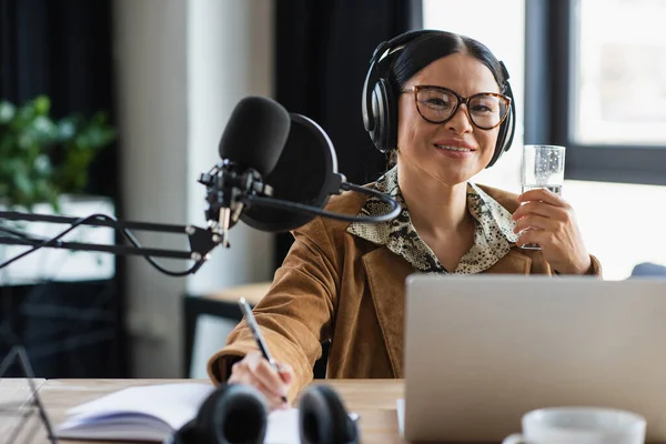 Позитивний азіатський радіоведучий в навушниках тримає склянку води і ручки біля блокнота в студії — стокове фото