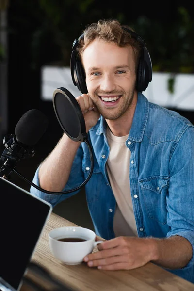 Anfitrião de rádio positivo em fones de ouvido segurando xícara de café em estúdio podcast — Fotografia de Stock