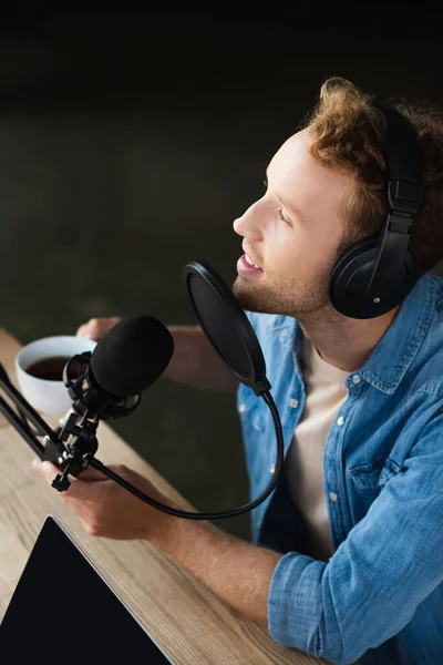 Vista de ángulo alto del presentador de radio positivo en auriculares que sostienen la taza de café en un estudio de podcast - foto de stock