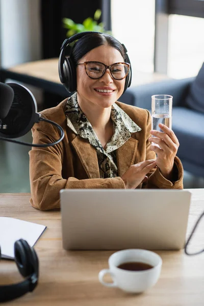 Веселий азіатський радіоведучий в навушниках тримає склянку води біля ноутбука в студії — стокове фото