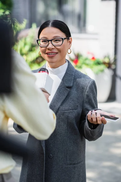 Cheerful asian businesswoman in glasses holding smartphone and giving interview near blurred reporter — Stock Photo