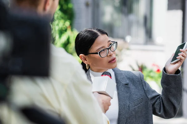 Азіатська бізнес-леді в окулярах тримає смартфон і дає інтерв'ю поблизу розмитого репортера — стокове фото