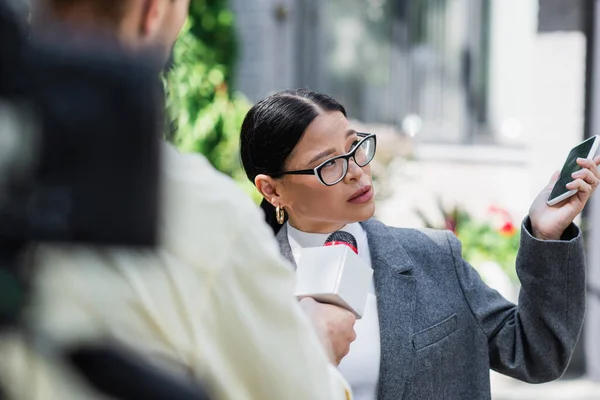 Asian businesswoman in glasses holding smartphone and giving interview near blurred reporter — Stock Photo