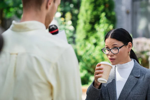 Asiatique femme d'affaires dans lunettes tenant tasse en papier et en regardant flou journaliste avec microphone — Photo de stock
