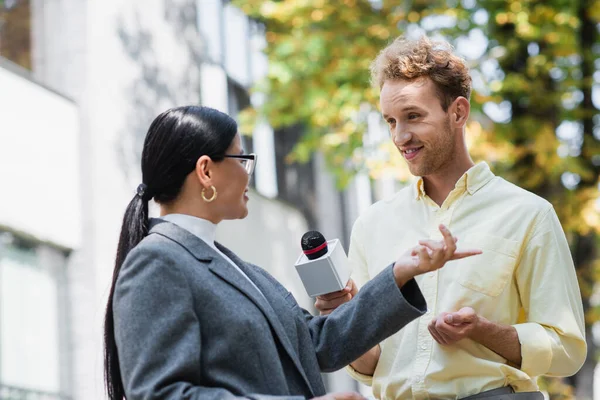 Cheerful reporter holding microphone near asian businesswoman pointing with finger while giving interview — Stock Photo