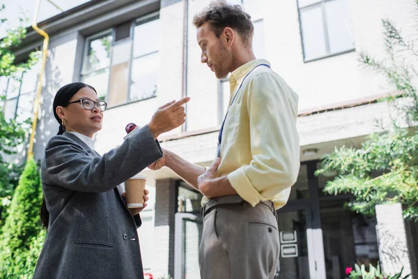Vue à faible angle du journaliste tenant un microphone près d'une femme d'affaires asiatique tenant une tasse en papier et pointant du doigt tout en donnant une interview — Photo de stock