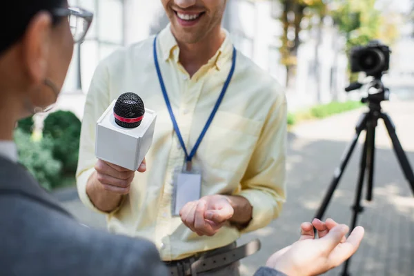 Cheerful reporter holding microphone near blurred businesswoman gesturing while giving interview — Stock Photo
