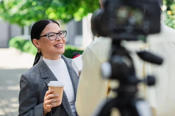 Borrosa reportero sosteniendo micrófono cerca feliz asiático mujer de negocios con papel taza dando entrevista - foto de stock