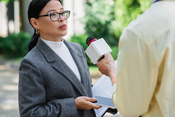 Репортер тримає мікрофон біля азіатської бізнес-леді в окулярах зі смартфоном — стокове фото