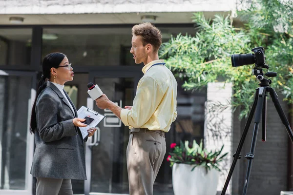 Side view of reporter holding microphone near asian businesswoman in glasses on street — Stock Photo