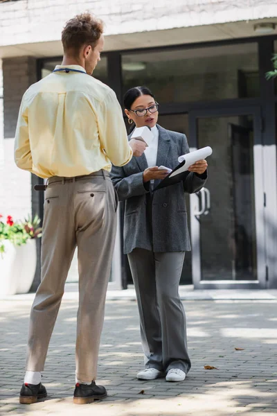 Full length of reporter taking interview with asian businesswoman in glasses holding smartphone and looking at clipboard on street — Stock Photo